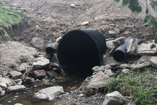 Large and small corrugated pipes emerging from a soil and rock embankment, with water flowing through the larger pipe.