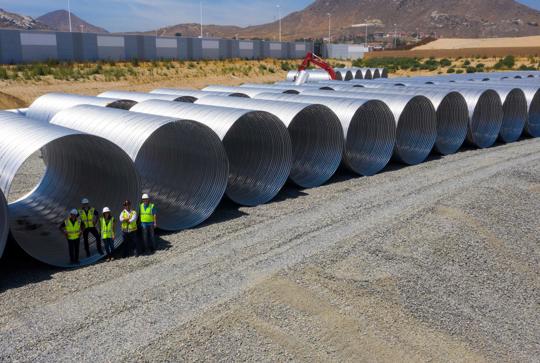Workers in safety vests stand beside large metal pipes stacked on a gravel lot, with a mountainous background.