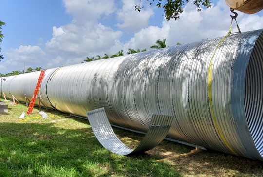 Large metal pipe being installed at a construction site with a crane, ladder, and visible cloudy sky.