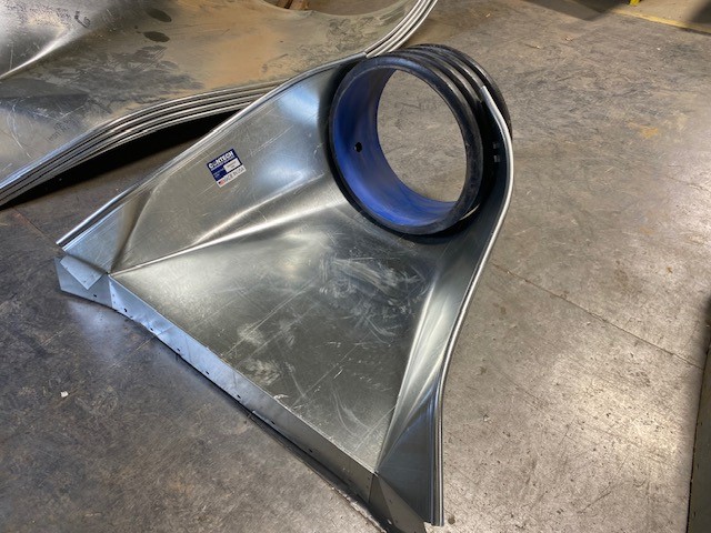 A large, angular metal duct piece with a circular opening sits on a workshop floor.
