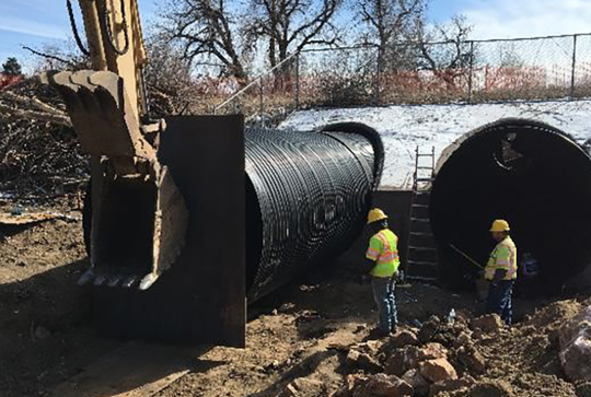 Construction workers in hard hats install large culvert pipes with the help of an excavator at a snowy worksite.