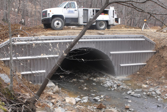 Construction site with a large corrugated metal culvert in a streambed, a utility truck parked on top, and bare trees around.