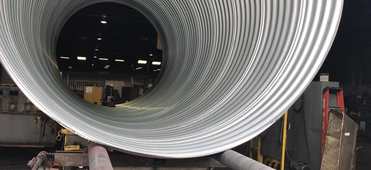 Large coiled metal sheet inside a manufacturing facility, with machinery and equipment visible in the background.
