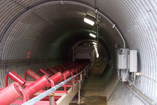 A long, corrugated metal tunnel with red mining conveyor equipment and electrical panels along the walls.