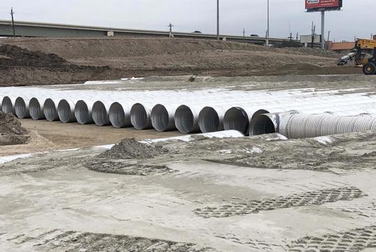 A row of large concrete pipes laid out in a construction area near a highway under a cloudy sky. A crane is visible nearby.