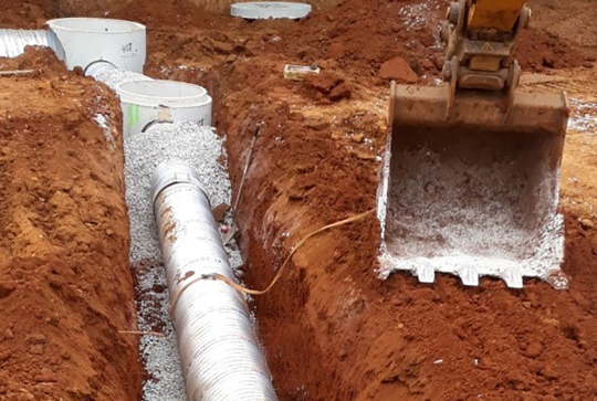 Trench with a large metal pipe and circular concrete structures, surrounded by dirt, alongside an excavator bucket.