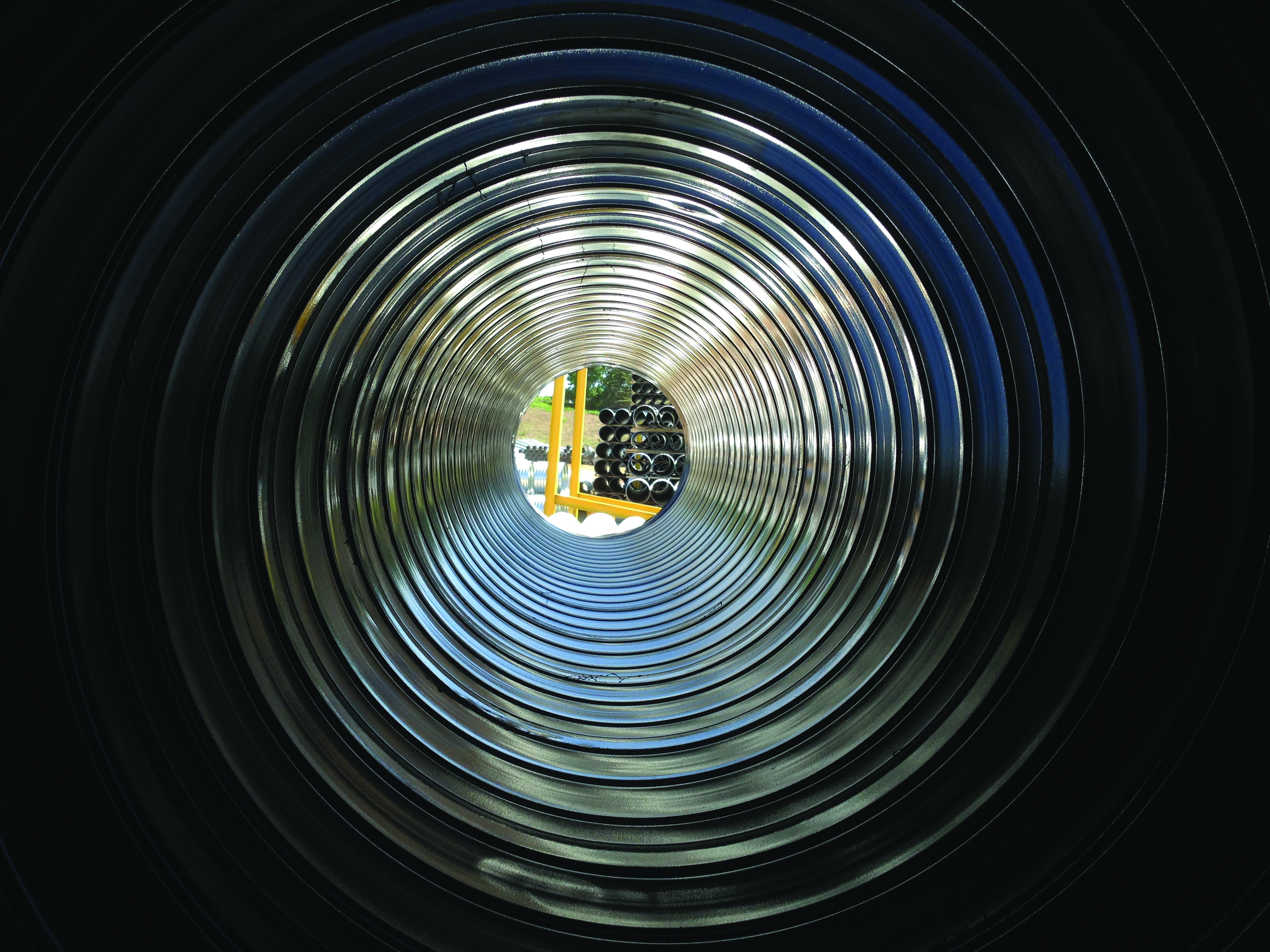 Inside of a large polymer-coated corrugated steel pipe, showing stacked pipes lined up outside in the background.