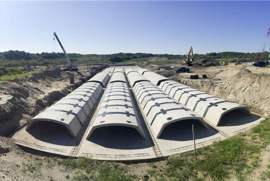 A row of large concrete structures under construction in an open field, with cranes and equipment in the background.