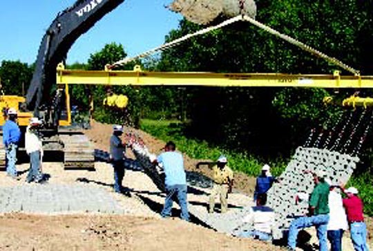 Construction crew uses a crane to lay concrete blocks at a site surrounded by trees.