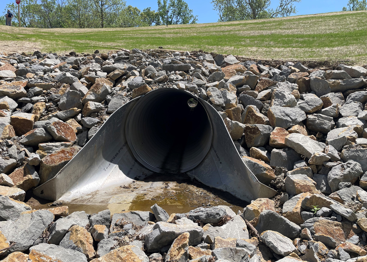 A large metal drainage pipe surrounded by rocks is embedded in a grassy embankment under a clear sky.