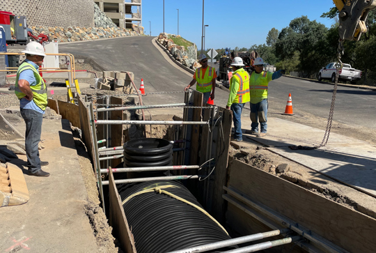 Workers in safety gear inspect equipment in a construction trench beside a road with traffic cones and a truck nearby.