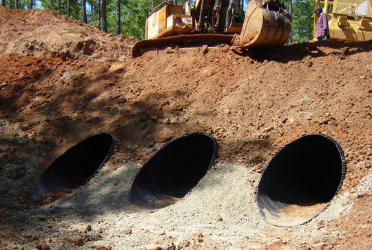 A construction site with three large culverts installed in the earth, with a bulldozer positioned above them.