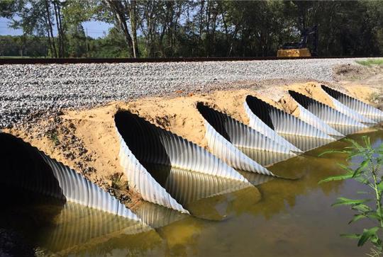 Six metal drainage culverts partially submerged in water beneath a gravel-covered railway track, surrounded by trees.