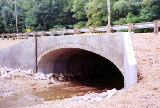 Concrete culvert under a road with a small stream flowing through, surrounded by grass and trees in the background.