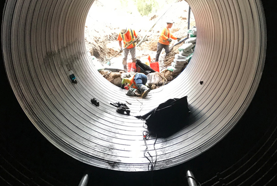 View through a large pipe under construction with three workers in safety gear, surrounded by construction materials and equipment.
