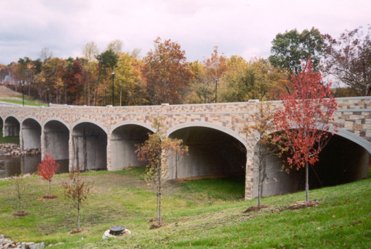 Stone bridge with arches over a river, surrounded by grassy area and trees with autumn foliage.