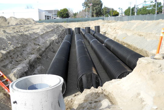 Large black drainage pipes installed in a trench at a construction site, surrounded by dirt and barriers.