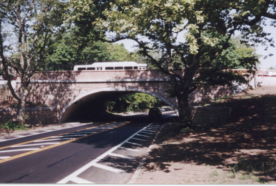 A stone bridge with a train passing over it spans a tree-lined road, with shadows cast on the pavement.