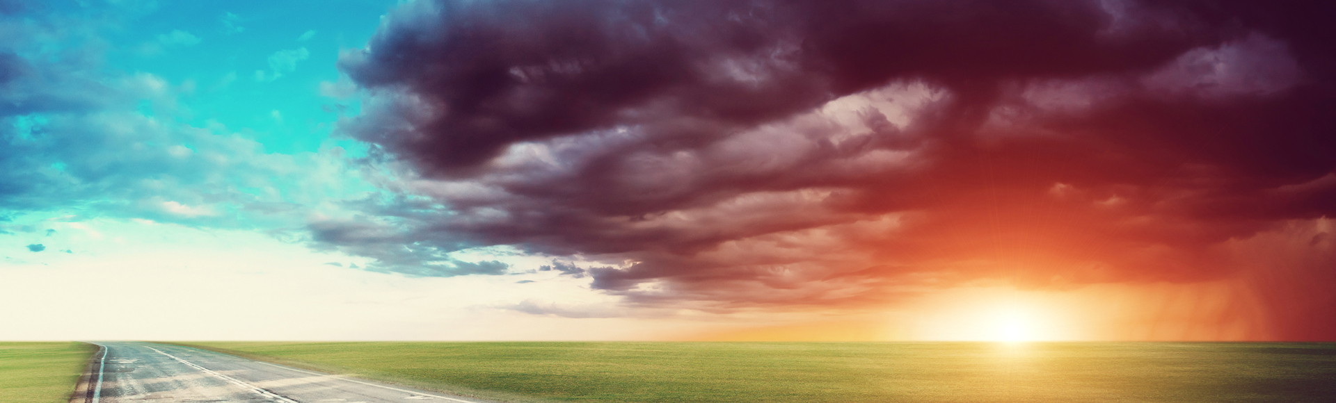 Empty road stretching into a vast landscape under a dramatic sky, split between blue and red hues, with the sun on the horizon.