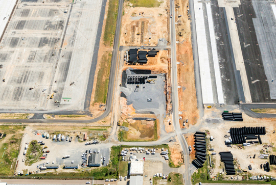 Aerial view of a large construction site with roads, machinery, and stacks of pipes alongside partially developed areas.