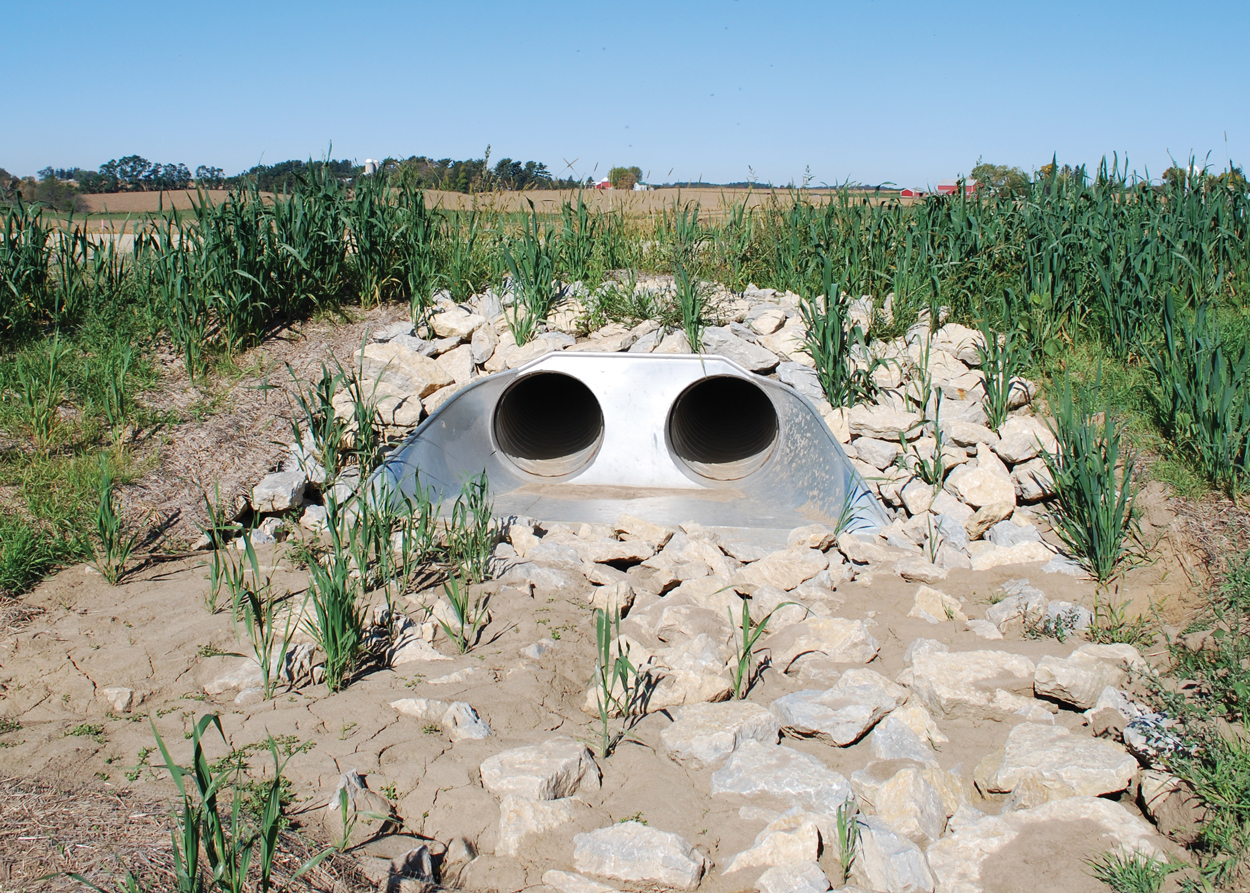 Twin metal drainage pipes surrounded by rocks and plants, set in a rural landscape under a clear sky.