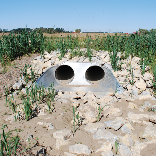 Twin metal drainage pipes surrounded by rocks and plants, set in a rural landscape under a clear sky.