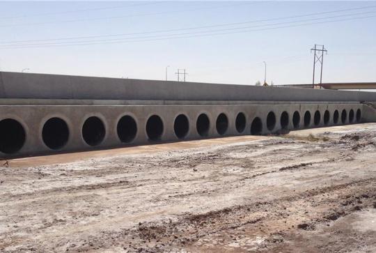 A concrete culvert with multiple circular openings for water flow is situated in a dry, dusty area under a clear sky.