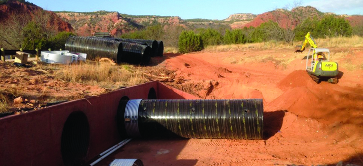 A construction site with large black pipes, excavated red soil, and a digger in a rocky landscape under a partly cloudy sky.
