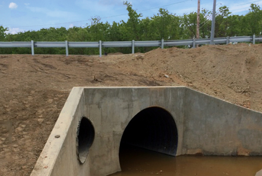 Concrete drainage culvert with two large circular openings, partially submerged in muddy water, surrounded by soil and greenery.
