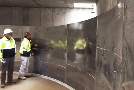 Two workers in safety gear examine a curved metallic structure inside an industrial setting.