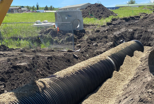 Construction site with a large black drainage pipe partially buried in gravel and dirt, surrounded by earth-moving equipment.