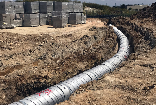 Trench with metal pipes laid underground in a construction site with stacked concrete blocks in the background.