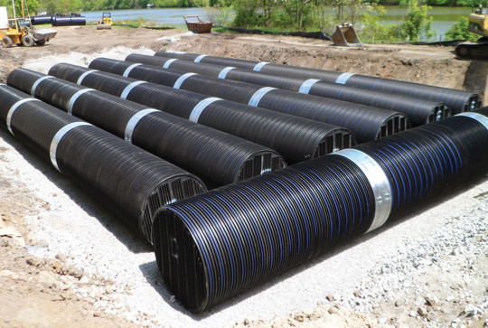 Large black drainage pipes arranged in parallel rows at a construction site with construction vehicles nearby.