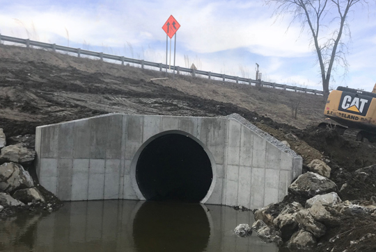 View of a concrete culvert tunnel near a body of water with an excavator and road construction sign in the background.