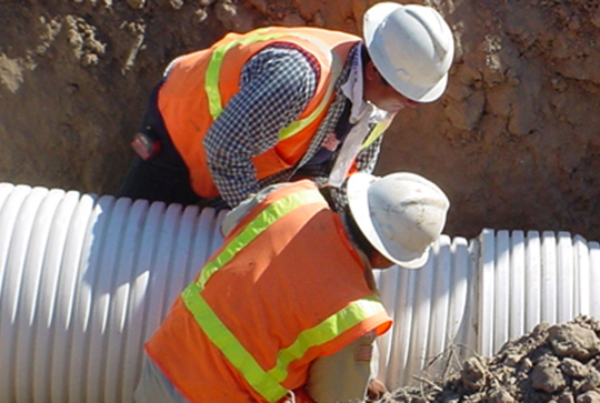 Two construction workers in safety gear install a large corrugated pipe in a dirt trench.